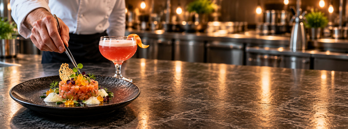 Chef che rifinisce una tartare di salmone con perle di agrumi, accanto a un cocktail al pompelmo